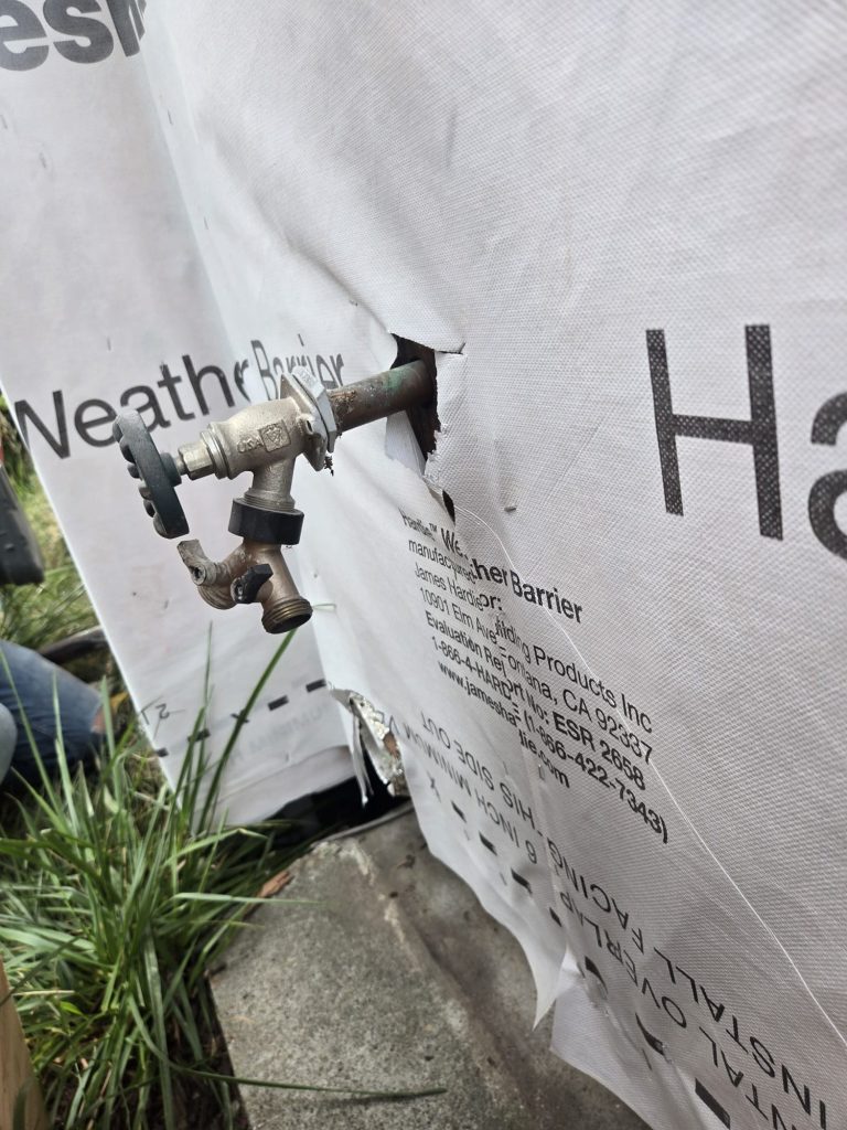 The existing brass hose bibb with a Y-splitter attached, sticking out through the weather barrier before replacement